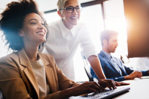 A group of people at a desk. Two women are on a computer and looking at the screen.