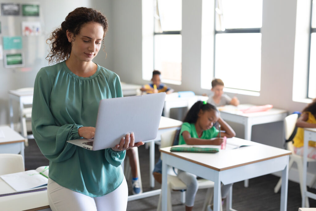 A teacher in a classroom is holding her laptop with young students sitting in their desks in the background.