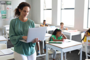 A teacher in a classroom is holding her laptop with young students sitting in their desks in the background.